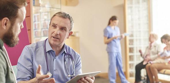 A male doctor sits next to a male patient in a waiting room while holding a digital tablet. , Credit: sturti via Getty Images A male doctor sits next to a male patient in a waiting room while holding a digital tablet. In the background a nurse chats to a family while holding digital tablet.