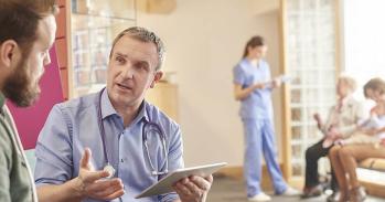 A male doctor sits next to a male patient in a waiting room while holding a digital tablet. In the background a nurse chats to a family while holding digital tablet.