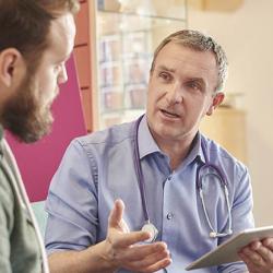 A male doctor sits next to a male patient in a waiting room while holding a digital tablet. In the background a nurse chats to a family while holding digital tablet.