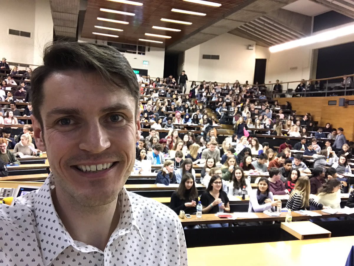 Ewan smiling with a raked-seating lecture hall full of students in the background.