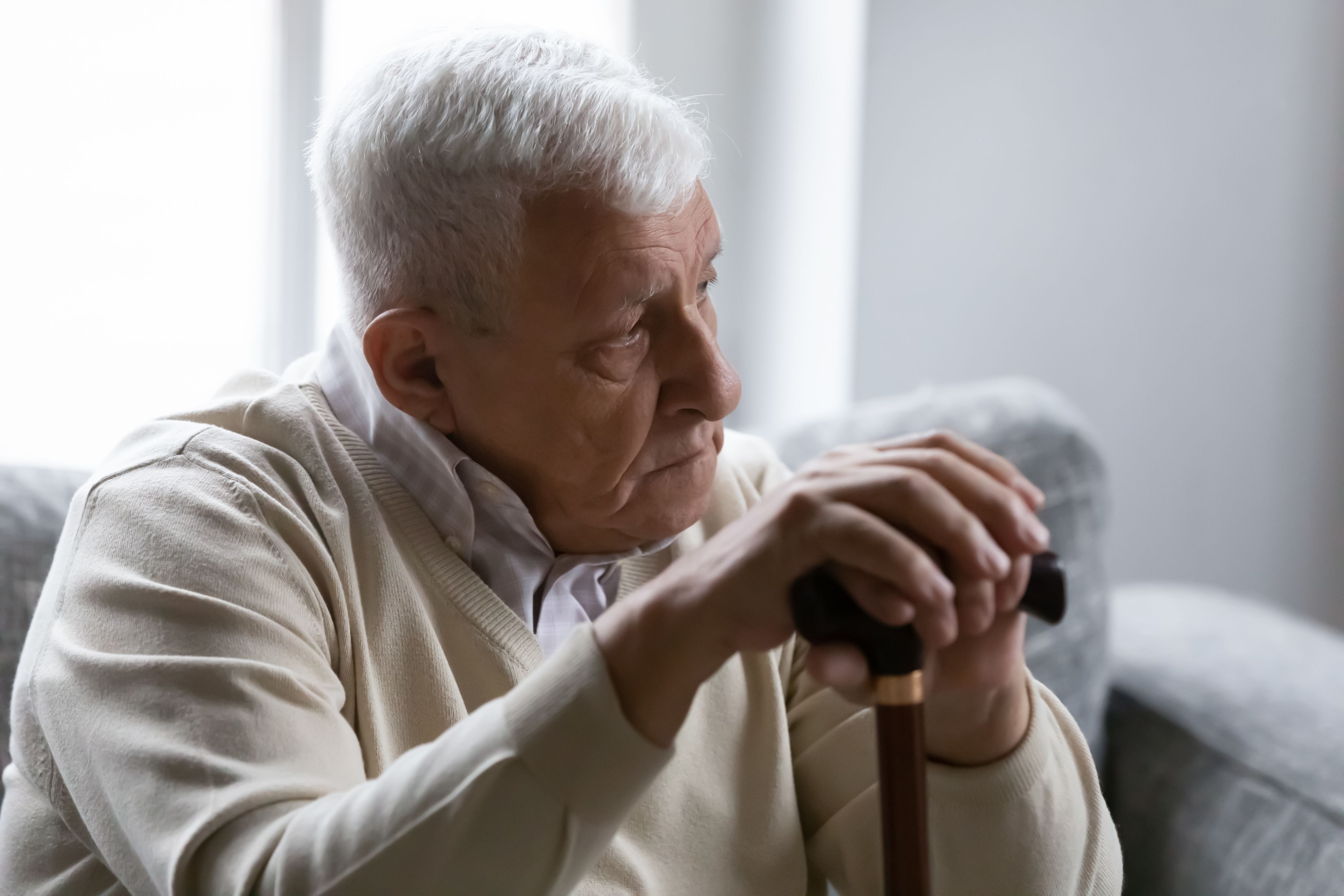 Old man holds cane sitting on couch looking at distance