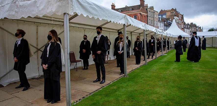 Graduands in academic dress for graduation standing in a socially distanced queue wearing masks