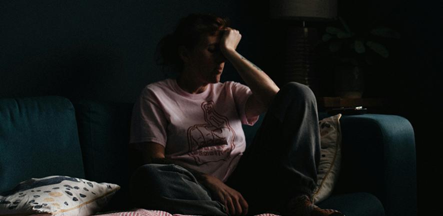 Woman sitting on sofa in the dark, placing a hand to her forehead.