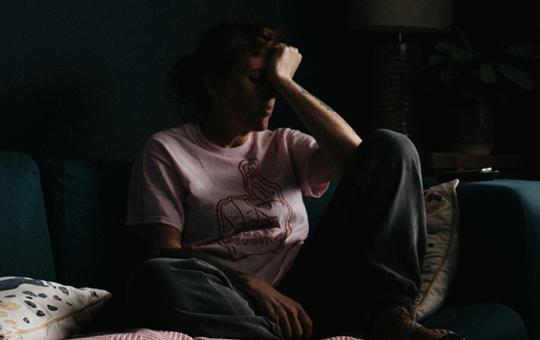 Woman sitting on sofa in the dark, placing a hand to her forehead.